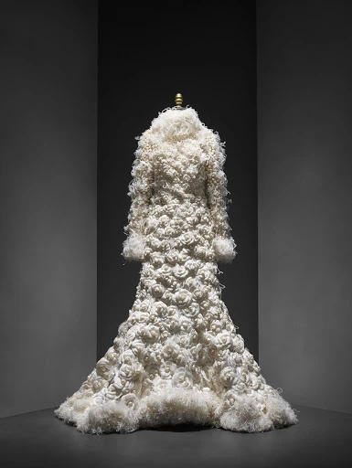 Full-length view of a couture ivory coat with textured rose appliqués, presented on a mannequin against a dark, graduated background.