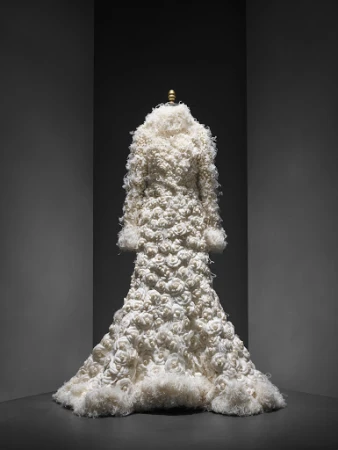 Full-length view of a couture ivory coat with textured rose appliqués, presented on a mannequin against a dark, graduated background.