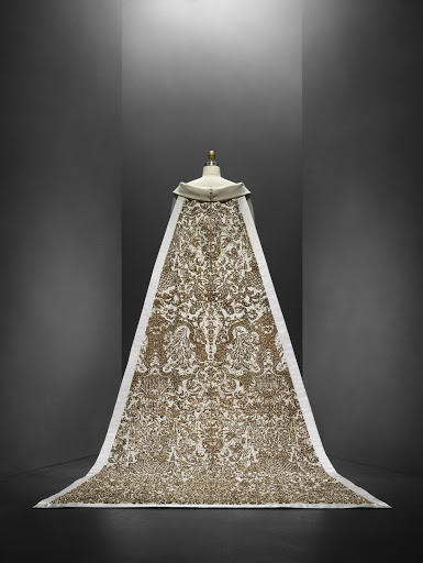 Ornate cream and gold patterned textile or gown draped over a mannequin, creating a dramatic display in a studio setting.