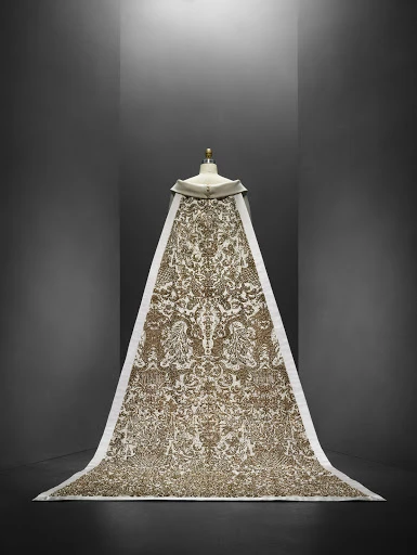 Ornate cream and gold patterned textile or gown draped over a mannequin, creating a dramatic display in a studio setting.