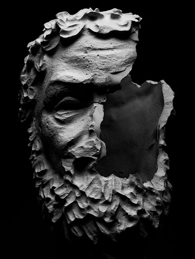 Close-up studio shot of a crumbling, damaged stone bust of a man with a beard against a stark black background, in monochrome.