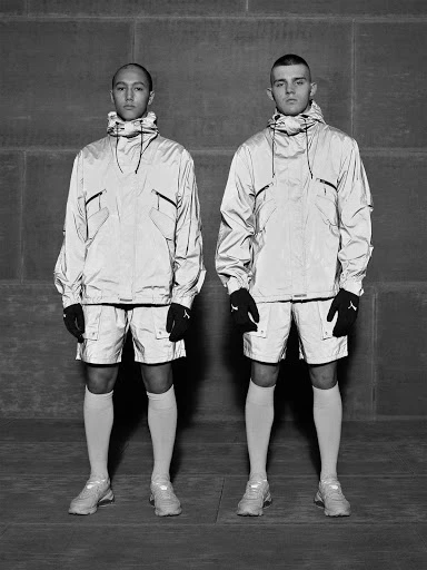 Two adult males pose in reflective athleisure wear against a textured grey background, showcasing modern sportswear and fashion.