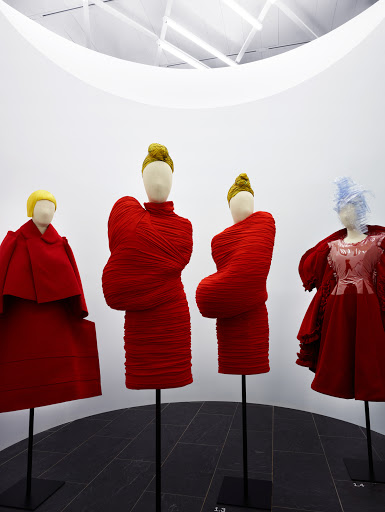 Fashion exhibit featuring four mannequins in red and pale-colored clothing, displayed in a minimalist art gallery setting with overhead lighting.