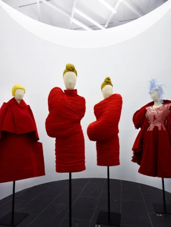 Fashion exhibit featuring four mannequins in red and pale-colored clothing, displayed in a minimalist art gallery setting with overhead lighting.