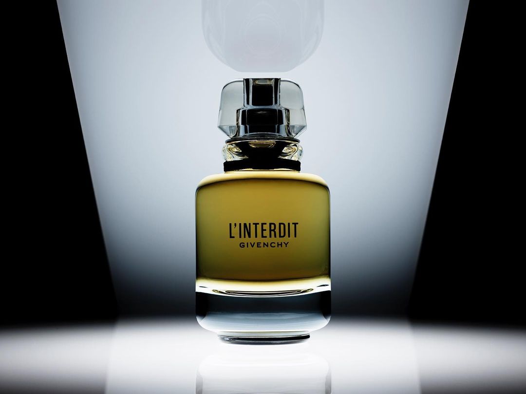 Close-up of a glass bottle of Givenchy L'Interdit perfume with a clear stopper against a black-and-white gradient background.