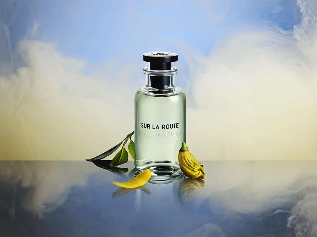 Still life shot of Louis Vuitton Sur La Route fragrance bottle with green leaves and yellow citrus fruit against a sky and smoke background.