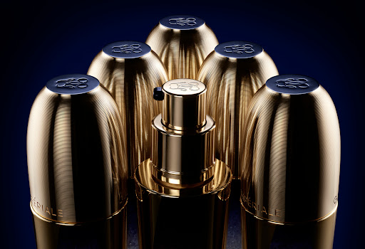 Close-up studio shot of gold-toned beauty serum and matching containers, set against a dark blue backdrop, promoting luxury skincare.
