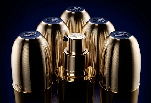 Close-up studio shot of gold-toned beauty serum and matching containers, set against a dark blue backdrop, promoting luxury skincare.