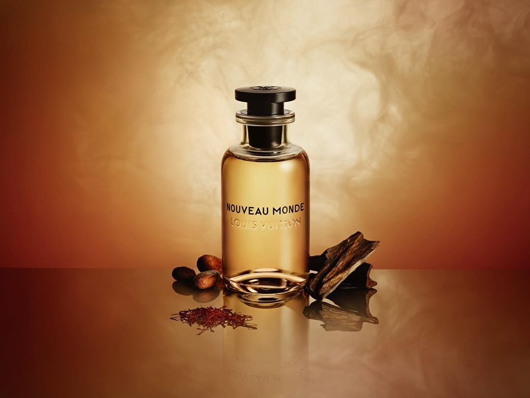Still life studio shot of a Louis Vuitton perfume bottle on a reflective surface with cocoa beans and wood pieces against a brown background.