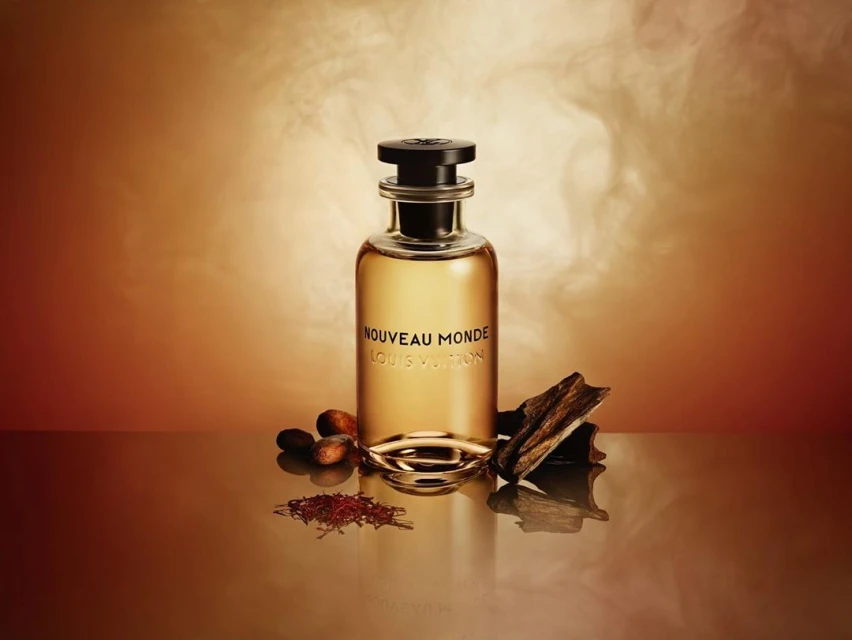 Still life studio shot of a Louis Vuitton perfume bottle on a reflective surface with cocoa beans and wood pieces against a brown background.