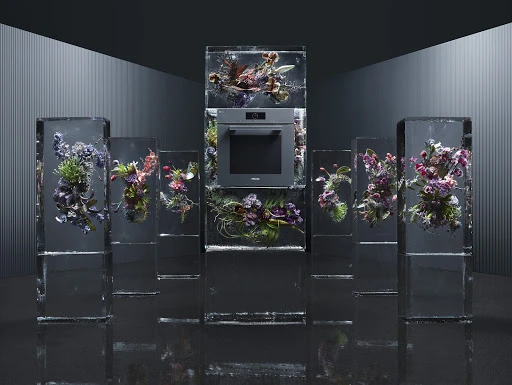 Modern kitchen appliance display featuring an oven encased in glass with floral arrangements, set against a dark, contemporary interior.