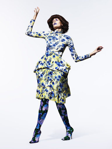 Full-length shot of a woman in a floral outfit striking a pose against a white background, showcasing a unique fashion aesthetic and bold makeup.