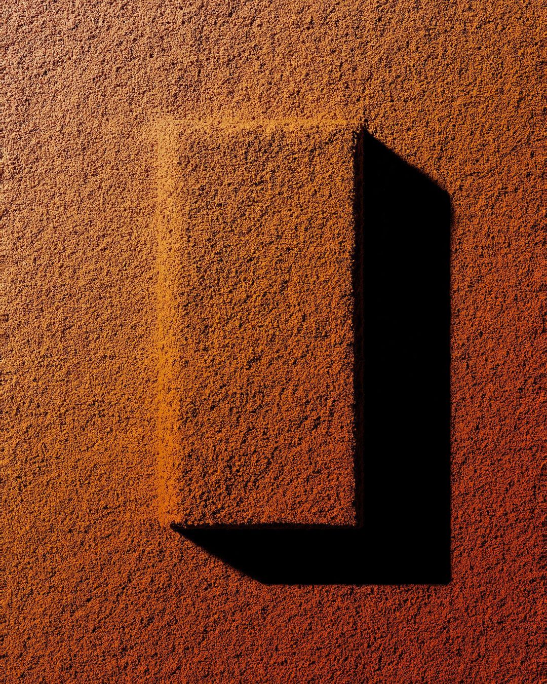 Abstract still life featuring a textured orange-brown surface with a raised rectangular impression, casting a stark black shadow.