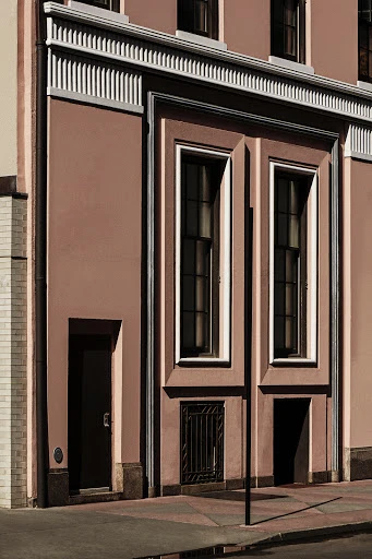 Facade of an Art Deco-style building with pink walls, symmetrical window placement, and geometric architectural details on a sunny day.