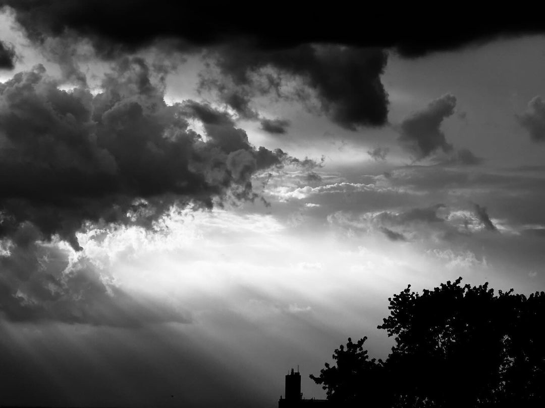 Dramatic black and white skyscape featuring storm clouds with sunlight breaking through, contrasted by silhouetted trees and distant architecture.