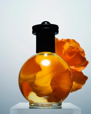 Round glass perfume bottle with a dark cap, filled with amber liquid, placed on a white pedestal with a vibrant orange flower behind it
