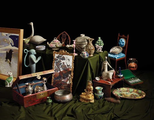 An assortment of antique objects and artifacts, including porcelain figures, vases, mirrors, and bowls, arranged on draped fabric against black backdrop.