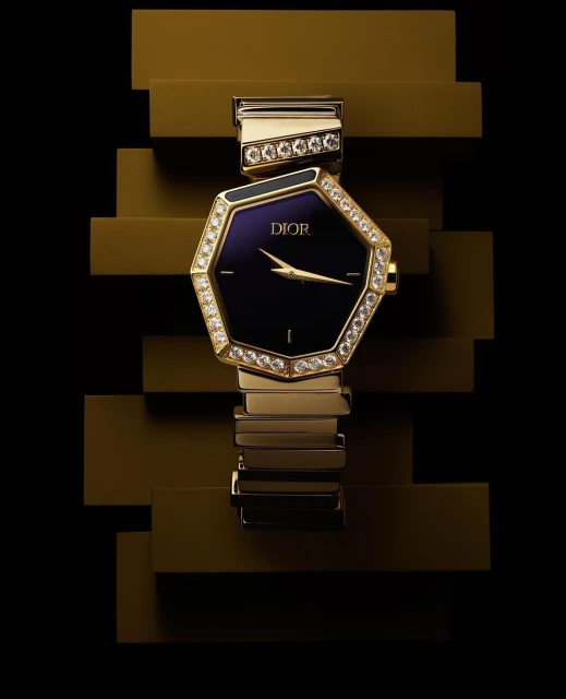 Octagonal Dior watch with a diamond bezel on a gold metal link bracelet against a stepped, glossy gold background and black background.