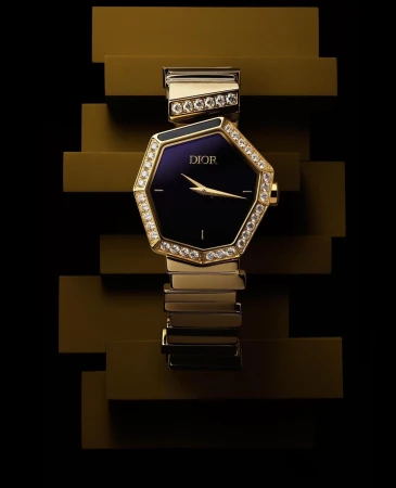 Octagonal Dior watch with a diamond bezel on a gold metal link bracelet against a stepped, glossy gold background and black background.