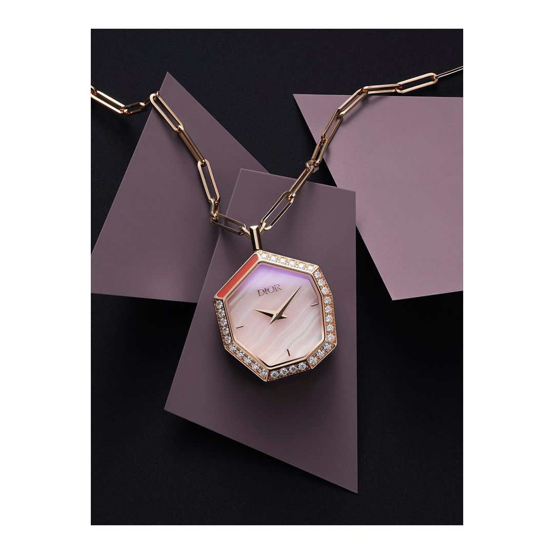 Elegant Dior watch pendant necklace with diamond bezel and chain, displayed against geometric lilac and black panels in a studio shot.