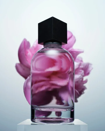 Glass perfume bottle with black cubic cap standing on a white pedestal before large pink flower petals