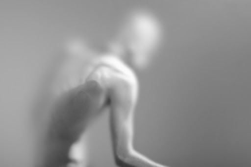 Monochromatic photograph of a blurred, shirtless adult male with arm raised in an indoor setting, focusing on form and light.