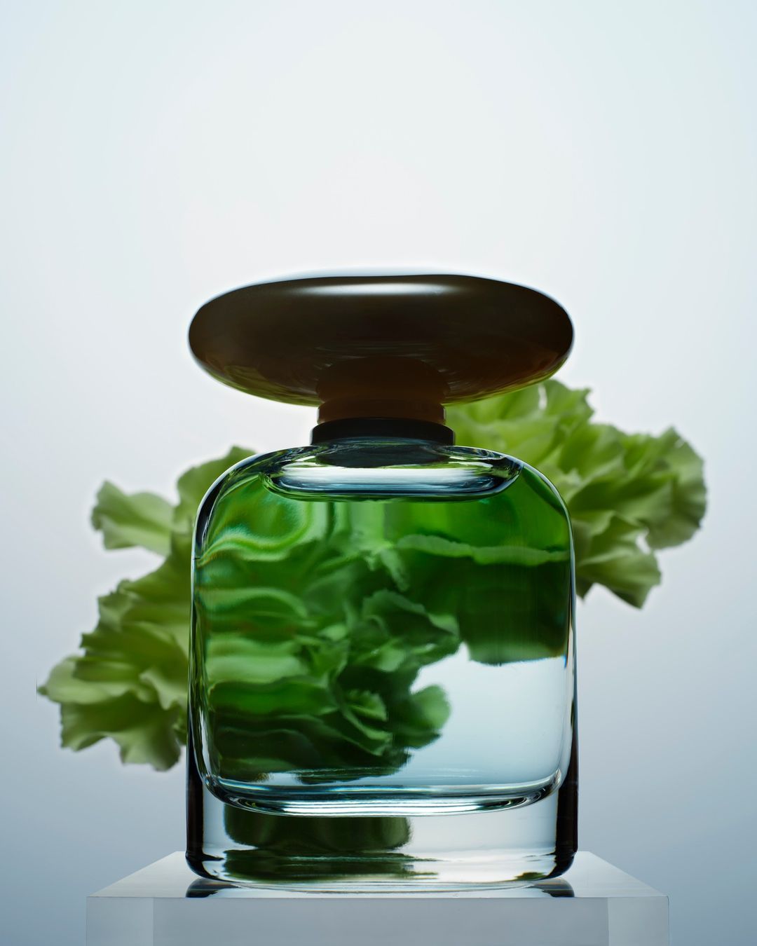 A glass perfume bottle with a brown cap sits on a white surface with soft green flowers behind the bottle in a beauty shot.