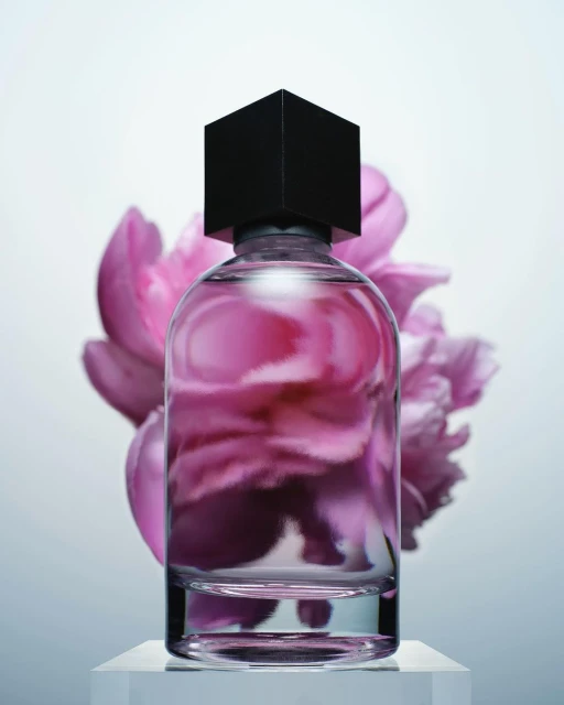 Studio shot of a clear perfume bottle with a black cube cap against a soft pink floral backdrop and white pedestal.