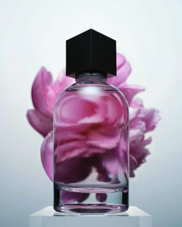Studio shot of a clear perfume bottle with a black cube cap against a soft pink floral backdrop and white pedestal.