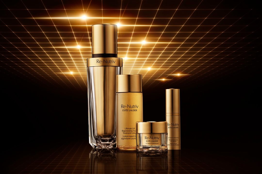 Five gold and clear glass Re-Nutriv Estee Lauder skincare product bottles on a dark surface, with a golden geometric pattern background.