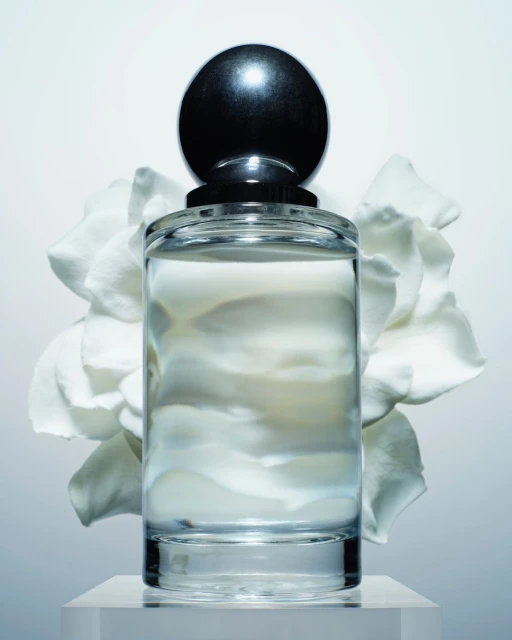 Product shot of a clear glass perfume bottle with a black spherical cap, set on a clear stand with a white flower in the background.