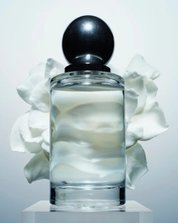 Product shot of a clear glass perfume bottle with a black spherical cap, set on a clear stand with a white flower in the background.