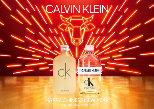 Two Calvin Klein fragrance bottles, CK One and CK Everyone, stand against a vibrant red backdrop with radiating orange neon lights.