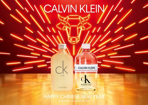 Two Calvin Klein fragrance bottles, CK One and CK Everyone, stand against a vibrant red backdrop with radiating orange neon lights.