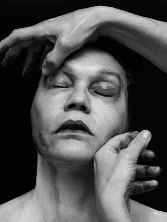 Close-up portrait in monochrome of a person with closed eyes as hands gently press against their face against a dark background.