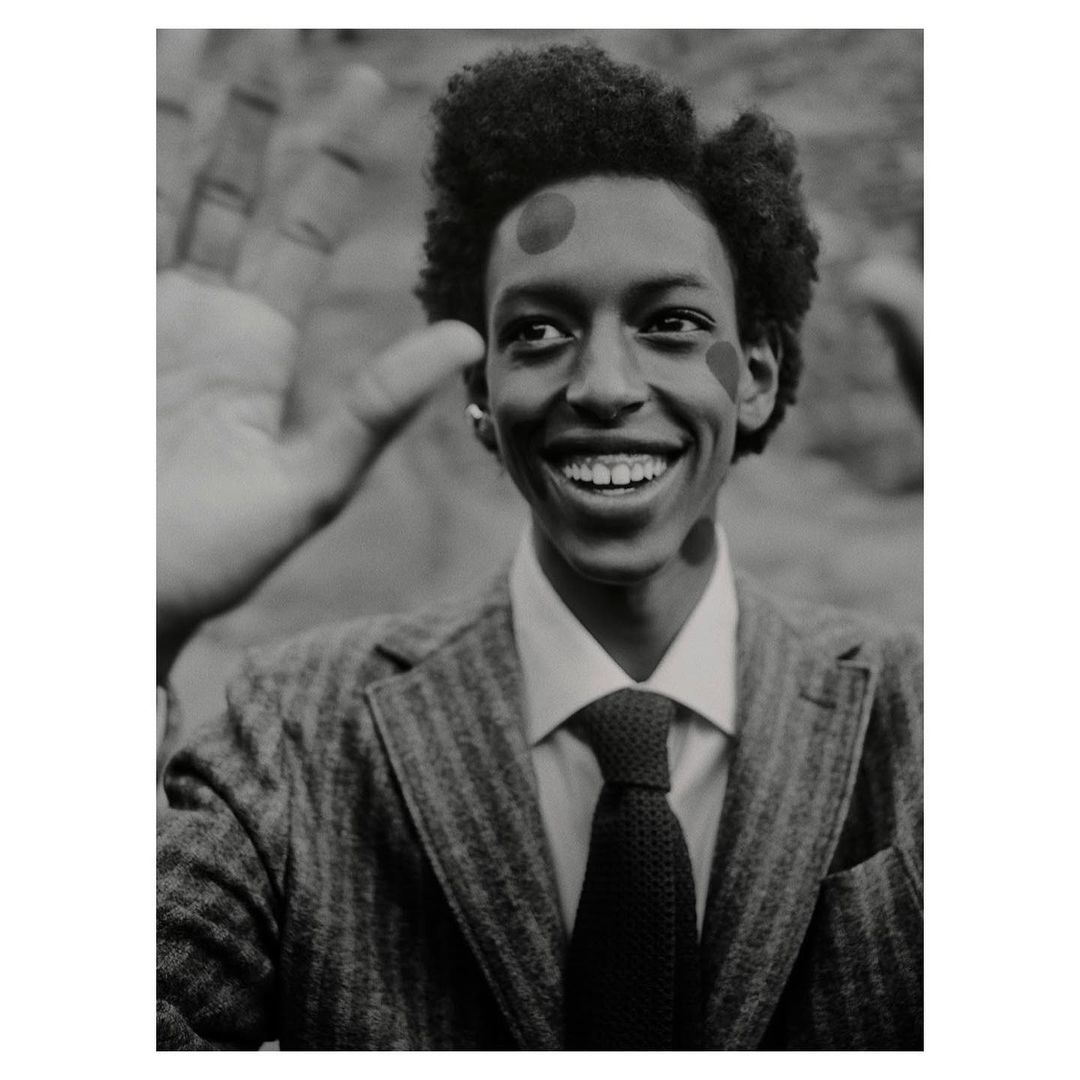 Smiling Black man in a suit with painted dots poses with a partial view of another person's hand in a monochrome studio shot.