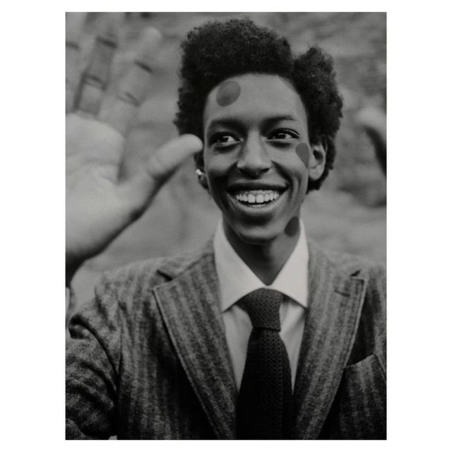 Smiling Black man in a suit with painted dots poses with a partial view of another person's hand in a monochrome studio shot.