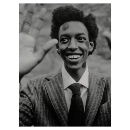 Smiling Black man in a suit with painted dots poses with a partial view of another person's hand in a monochrome studio shot.