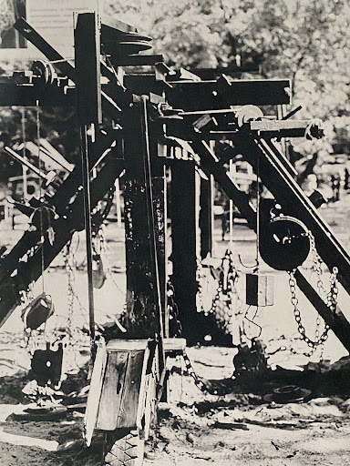 Monochrome image of a wooden playground structure with chains and pulleys, set in an outdoor park area.