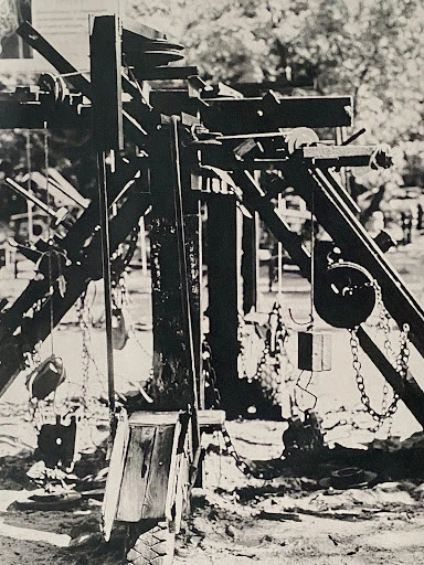 Monochrome image of a wooden playground structure with chains and pulleys, set in an outdoor park area.