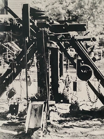 Monochrome image of a wooden playground structure with chains and pulleys, set in an outdoor park area.