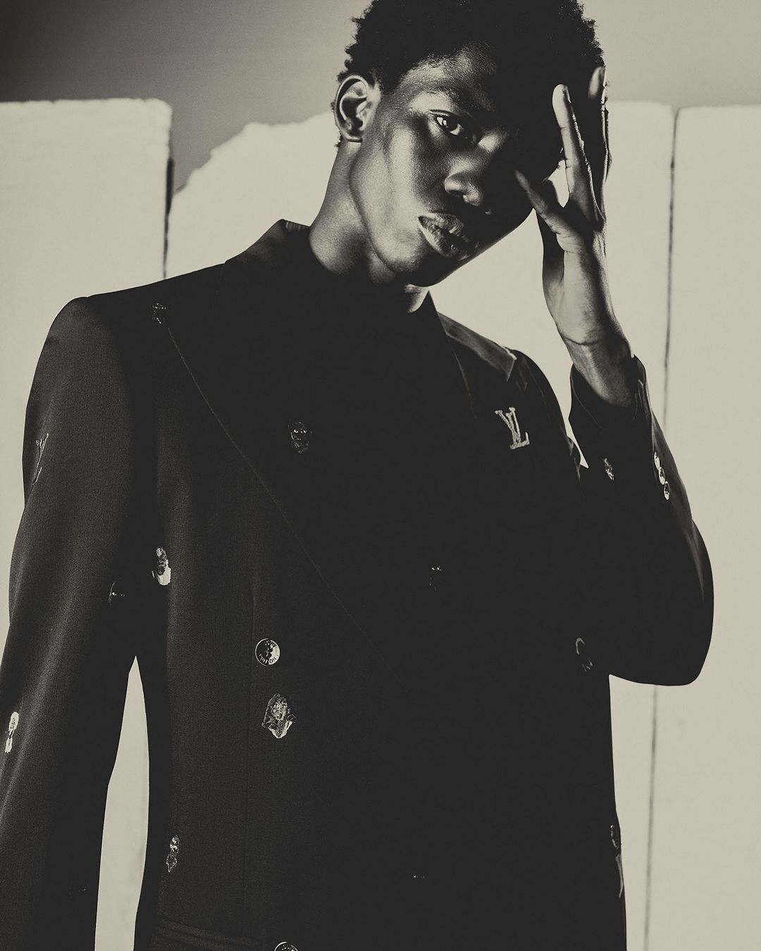 Portrait of a young adult Black male model in a stylish black blazer with decorative buttons and a Louis Vuitton logo against a grey background.