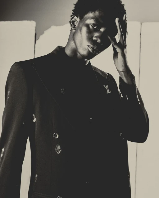 Portrait of a young adult Black male model in a stylish black blazer with decorative buttons and a Louis Vuitton logo against a grey background.