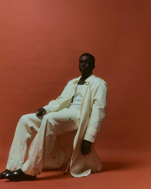 Man in a cream-colored trench coat sits in a photo studio against an orange backdrop, wearing a white tank top and cream trousers.