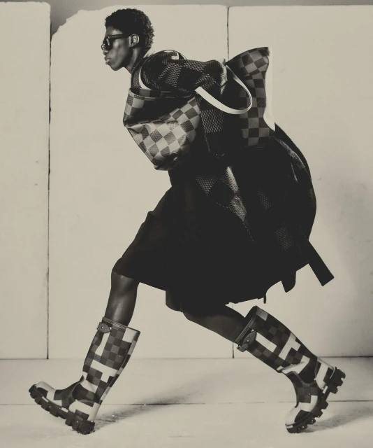 Monochrome fashion shot of a Black model striding in a checkered-pattern jacket, shorts, boots, and bag against a neutral backdrop.
