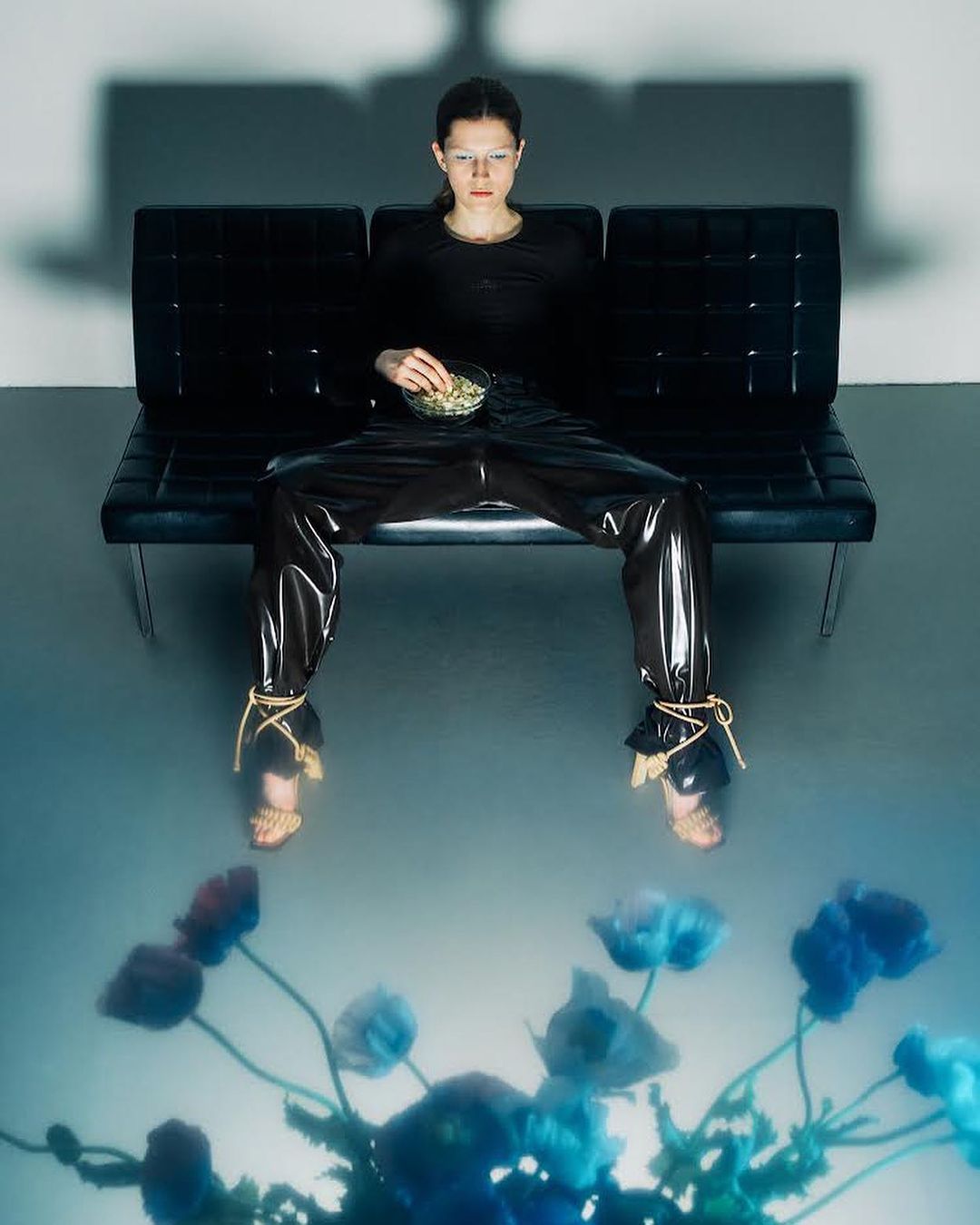 Woman in glossy pants on a black couch with a bowl of food, reflected in a flower pattern against a grey studio backdrop.