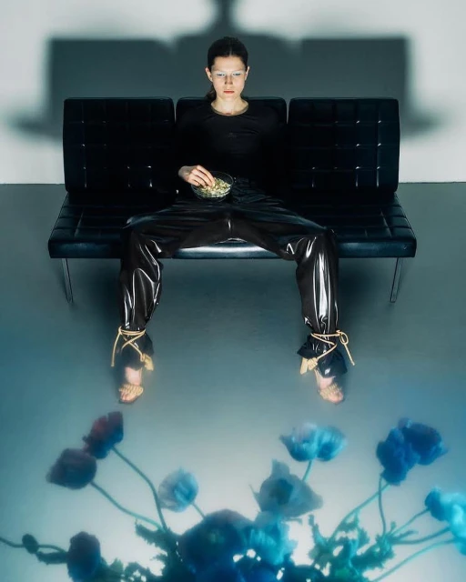 Woman in glossy pants on a black couch with a bowl of food, reflected in a flower pattern against a grey studio backdrop.