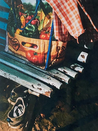 A reusable shopping bag with a vibrant vegetable print rests on a weathered green wooden bench with sneakers underneath.
