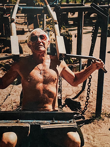 An elderly man with sunglasses is seen working out on a chest press machine outdoors in natural lighting, with foliage in the background.