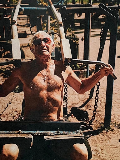 An elderly man with sunglasses is seen working out on a chest press machine outdoors in natural lighting, with foliage in the background.